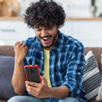 Happy Indian male receives online gift feeling joy sitting at home on the couch. Excited young man holding mobile phone reading good news winning lottery game