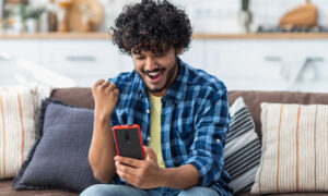 Happy Indian male receives online gift feeling joy sitting at home on the couch. Excited young man holding mobile phone reading good news winning lottery game